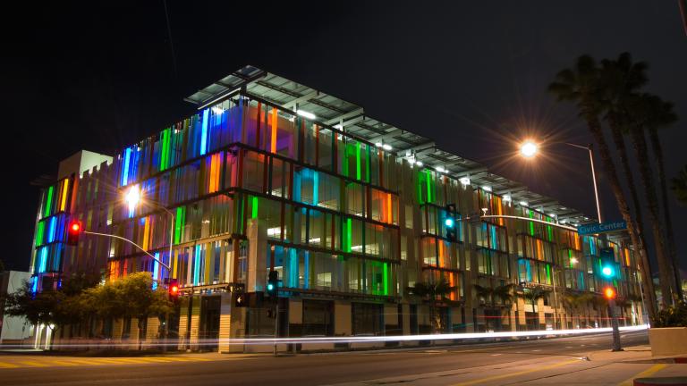 The exterior of a modern, glass-clad parking garage, lit up at night with different colors shining out.