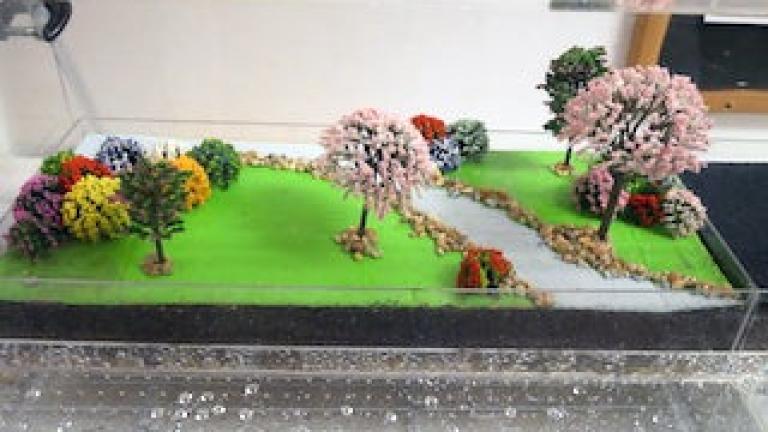 Photo of a diorama, with trees, grassy areas, and a stream, demonstrating water runoff and absorption.