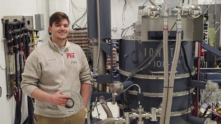 MIT graduate student Daniel Korsun holds a reel of the high-temperature superconducting tape that has been the focus of his research, as he stands beside the cyclotron he uses in his experiments. 