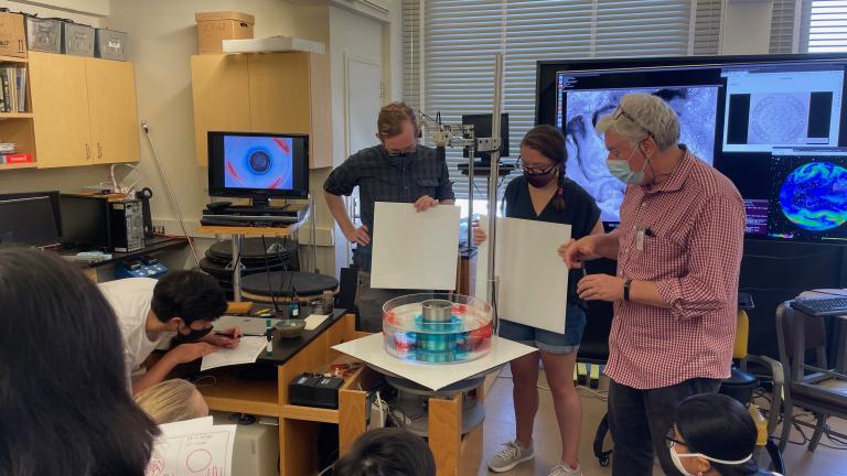 Three people, two of whom are holding large, white square boards, are looking down at a circular device filled with light blue and red dye.