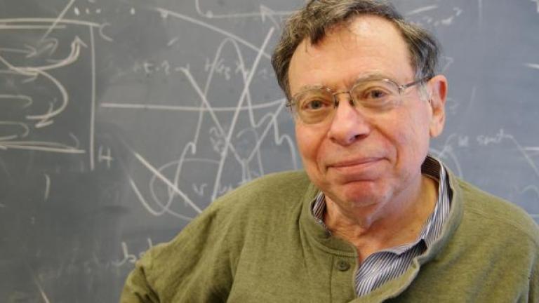 A portrait of Carl Wunsch standing in front of a blackboard with white chalk math drawings in the background.