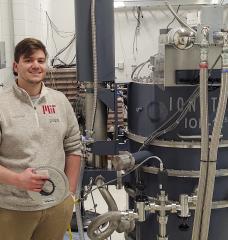 MIT graduate student Daniel Korsun holds a reel of the high-temperature superconducting tape that has been the focus of his research, as he stands beside the cyclotron he uses in his experiments. 