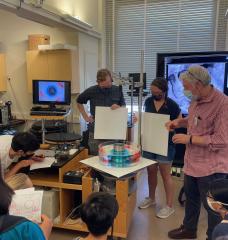 Three people, two of whom are holding large, white square boards, are looking down at a circular device filled with light blue and red dye.