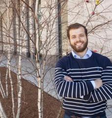 Photo of Theo Mouratidis, crossed arms, standing on MIT campus near birch trees and building